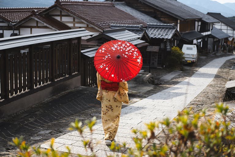 Best time to visit Japan Woman in kimono with red umbrella walking through a traditional Japanese village street.