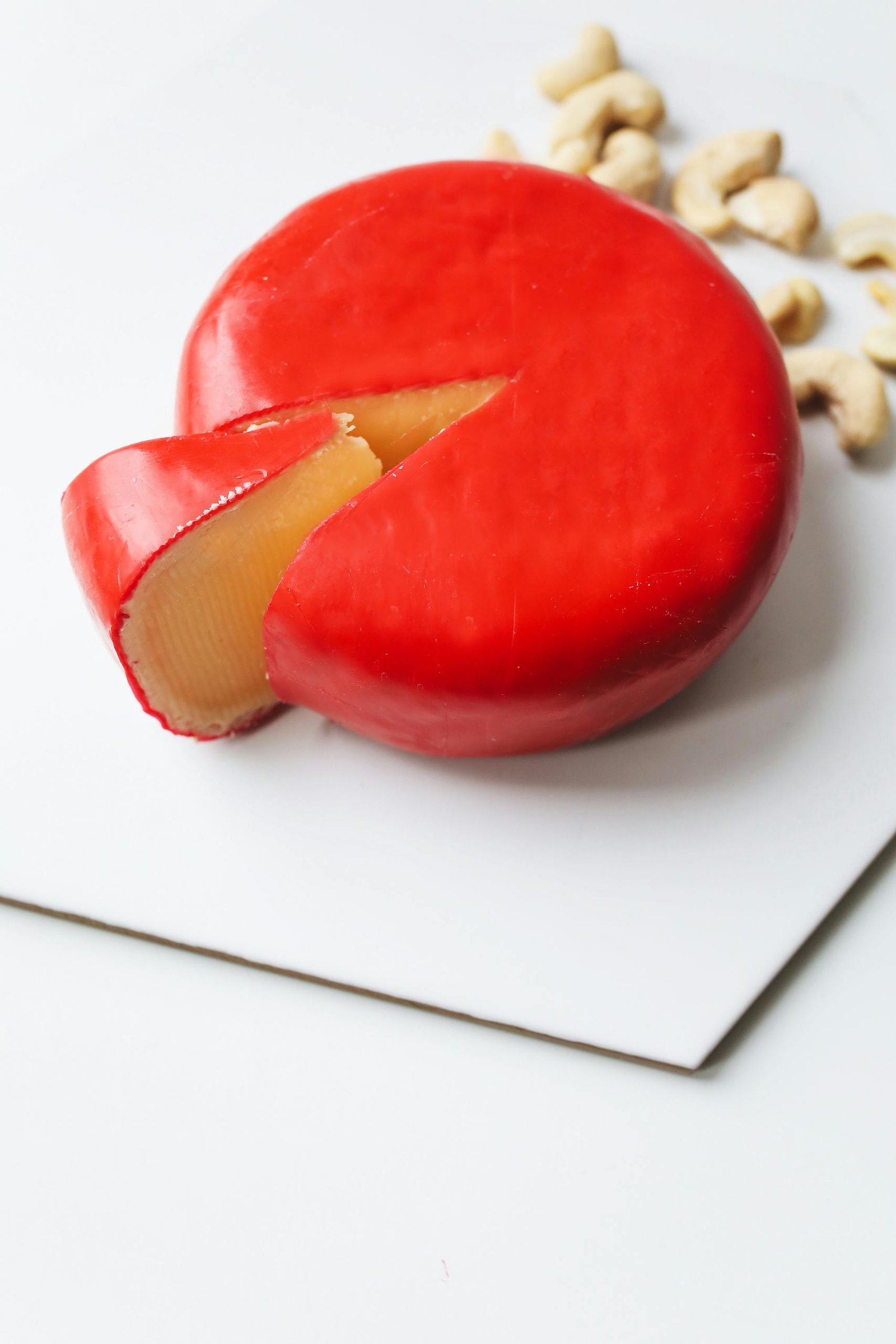 A close-up of Gouda cheese with a wedge cut, accompanied by cashew nuts on a white surface.