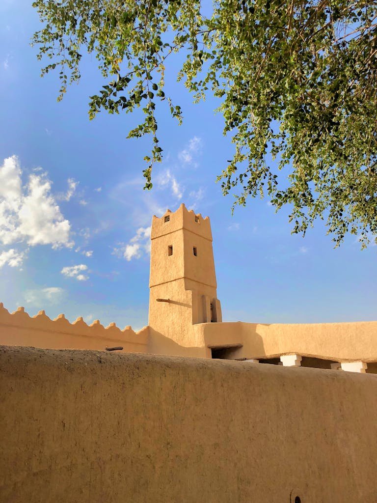 A historic tower in Diriyah, Saudi Arabia, framed by tree branches under a bright blue sky.