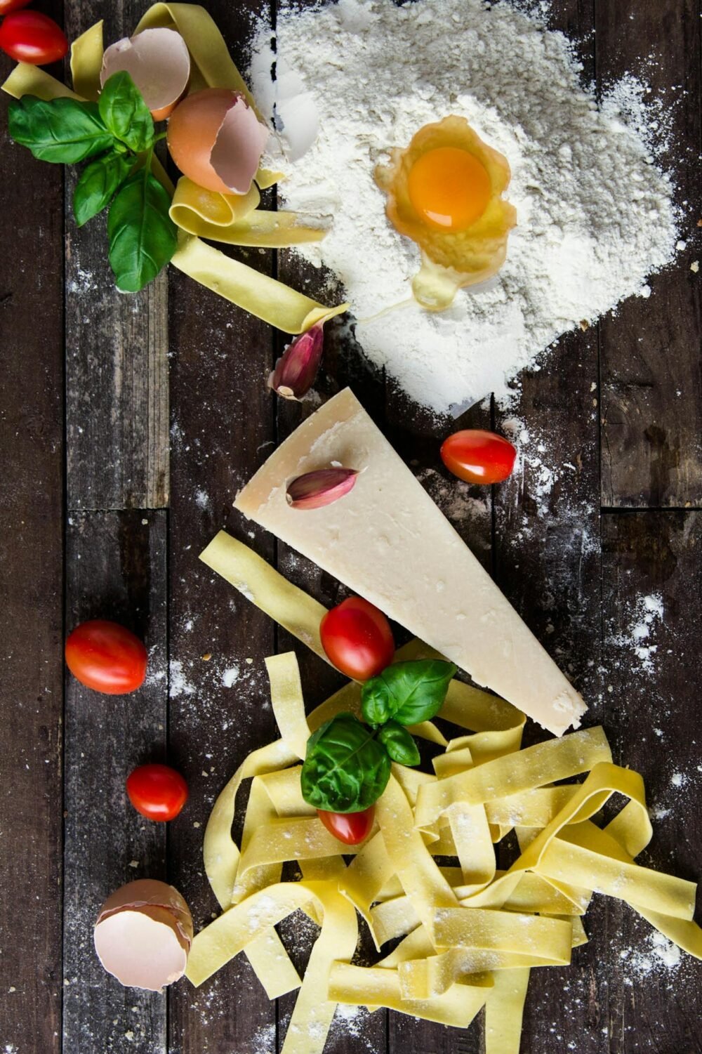 Top view of fresh pasta ingredients including flour, egg, parmesan, cherry tomatoes, and basil on a rustic wooden surface.