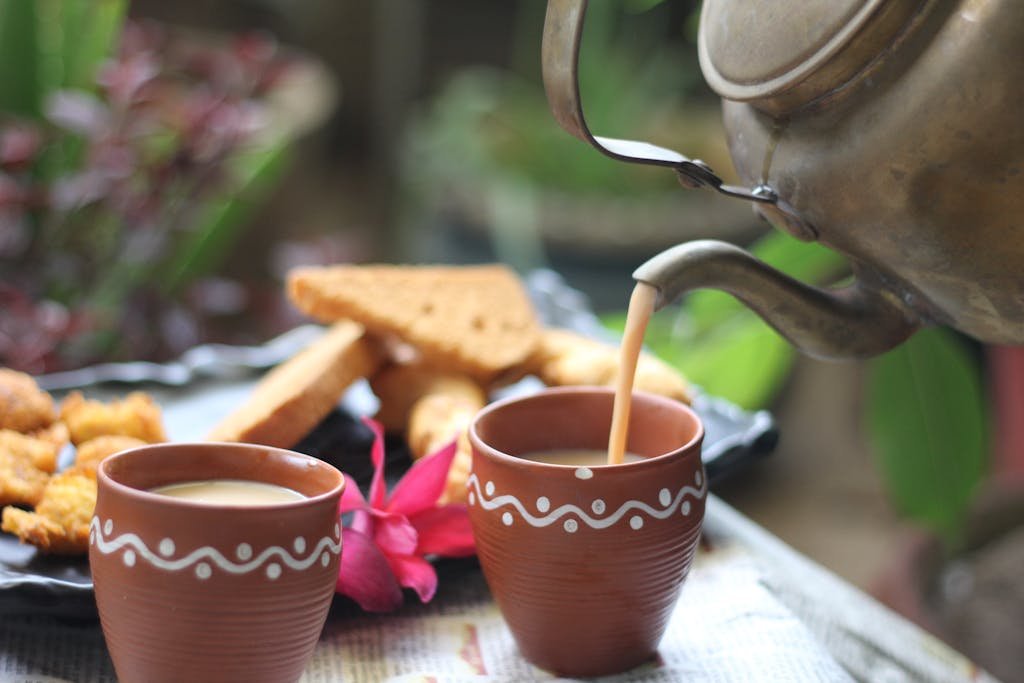 A warm cup of tea being poured from a kettle, surrounded by snacks and a flower.