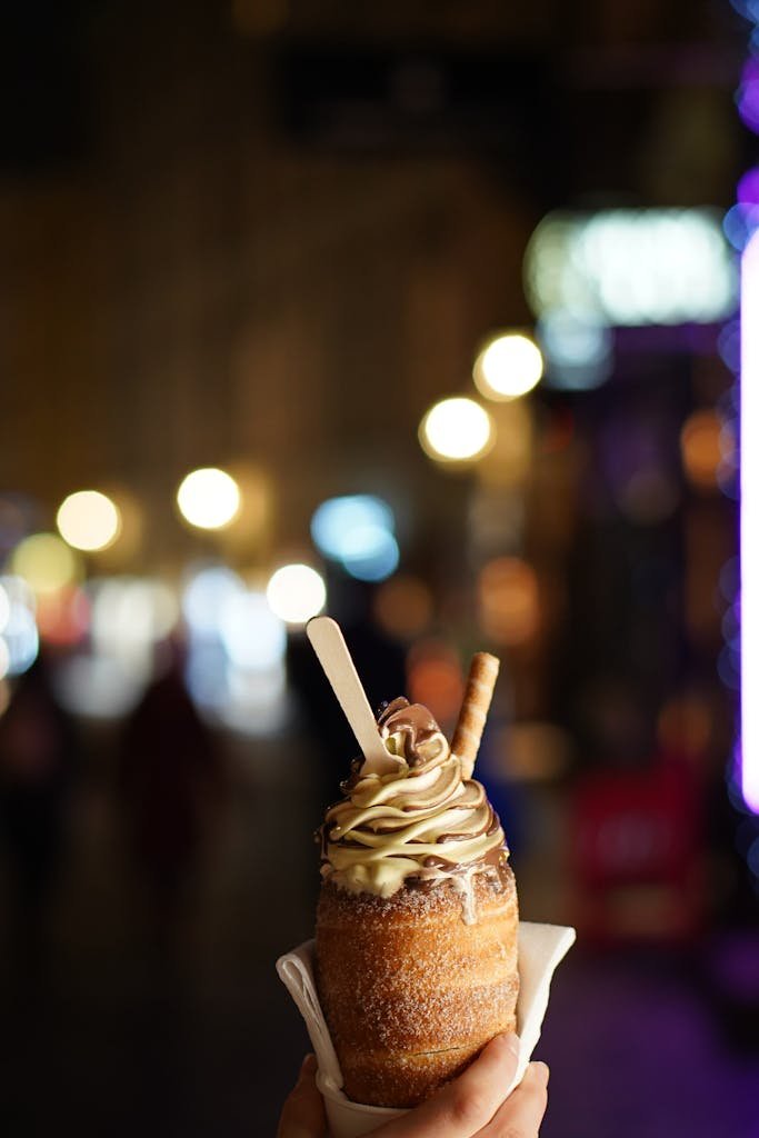 Hand holding a Trdelník ice cream dessert with lights blurred in Prague's night ambiance.