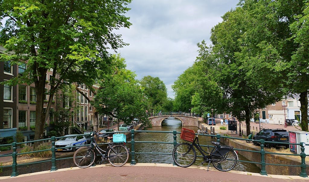 9 Ways of Getting Around Amsterdam Like a Local 9 Scenic view of bicycles on a bridge overlooking a canal in Amsterdam, lined with trees and historic buildings.