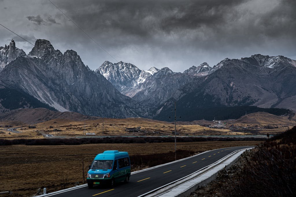 Blue van travels along a scenic mountain road with snow-dusted peaks under a cloudy sky best travel quotes