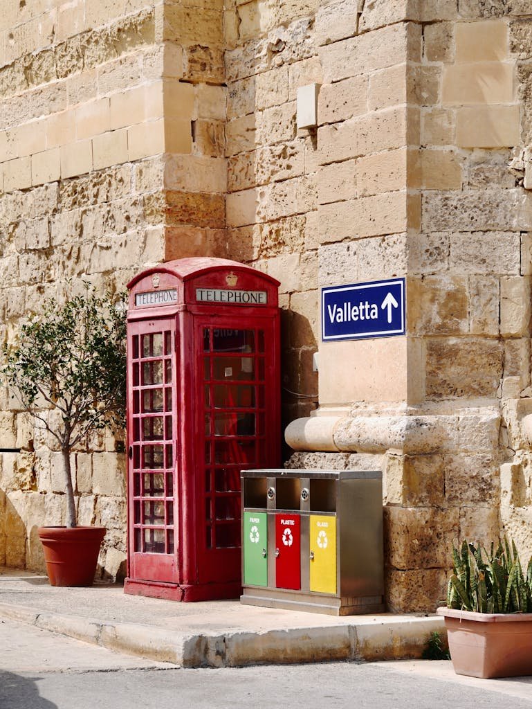 Classic red phone booth beside recycling bins in Valletta, Malta, showcasing traditional architecture.