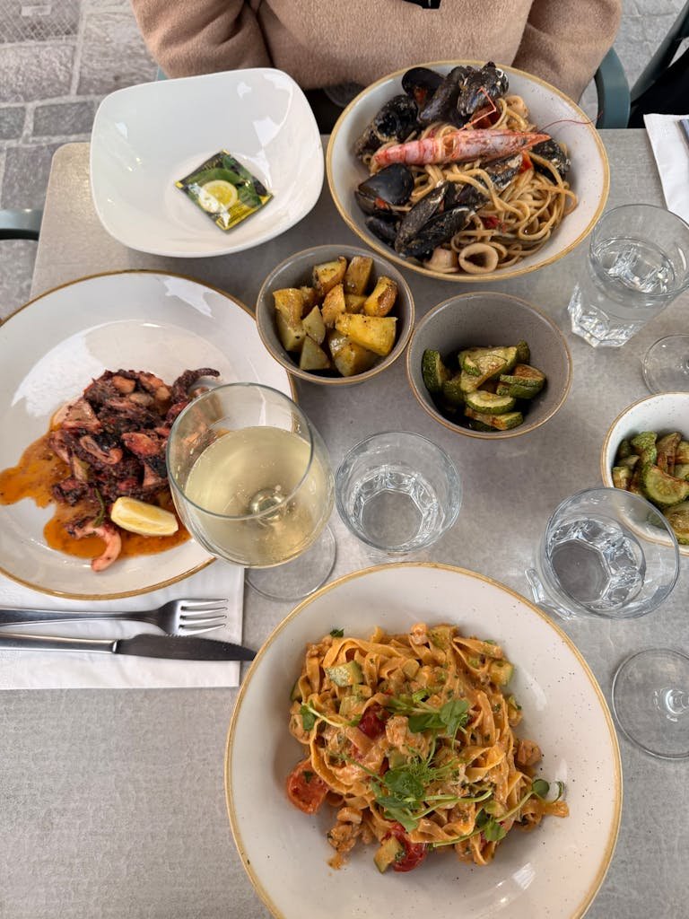 Delicious gourmet meal featuring seafood pasta, roasted vegetables, and wine in a Maltese cafe setting.