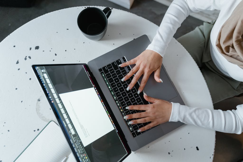 Overhead view of a woman's hands typing on a laptop at a white round table with coffee cup.