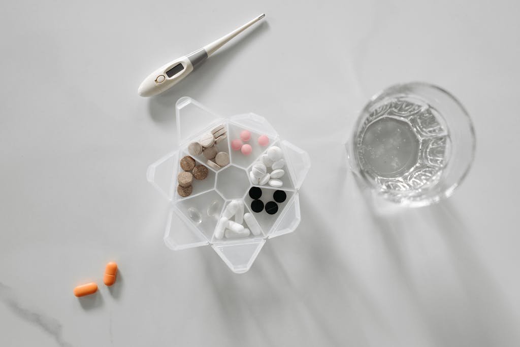 Overhead view of assorted medications with glass of water and digital thermometer on white background.