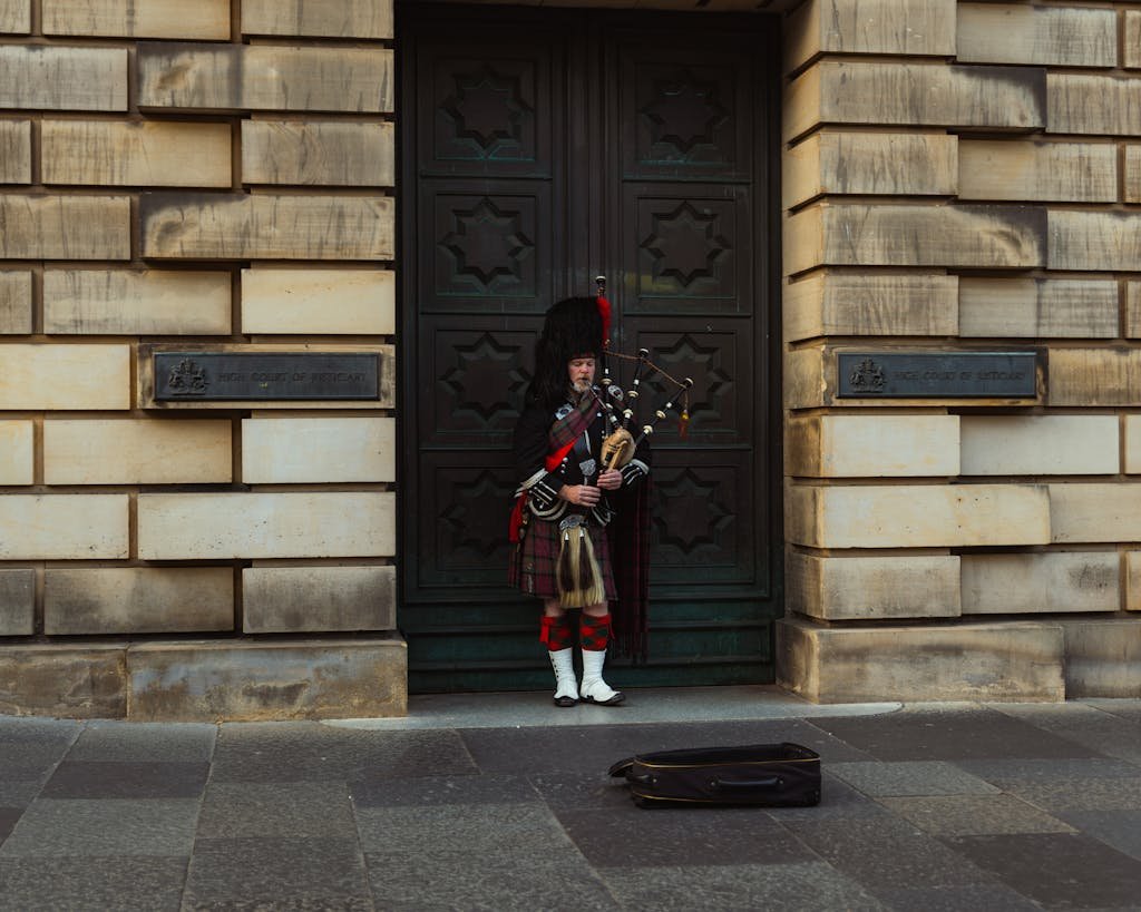 A bagpiper in traditional attire plays outside a historic building in Edinburgh, Scotland.