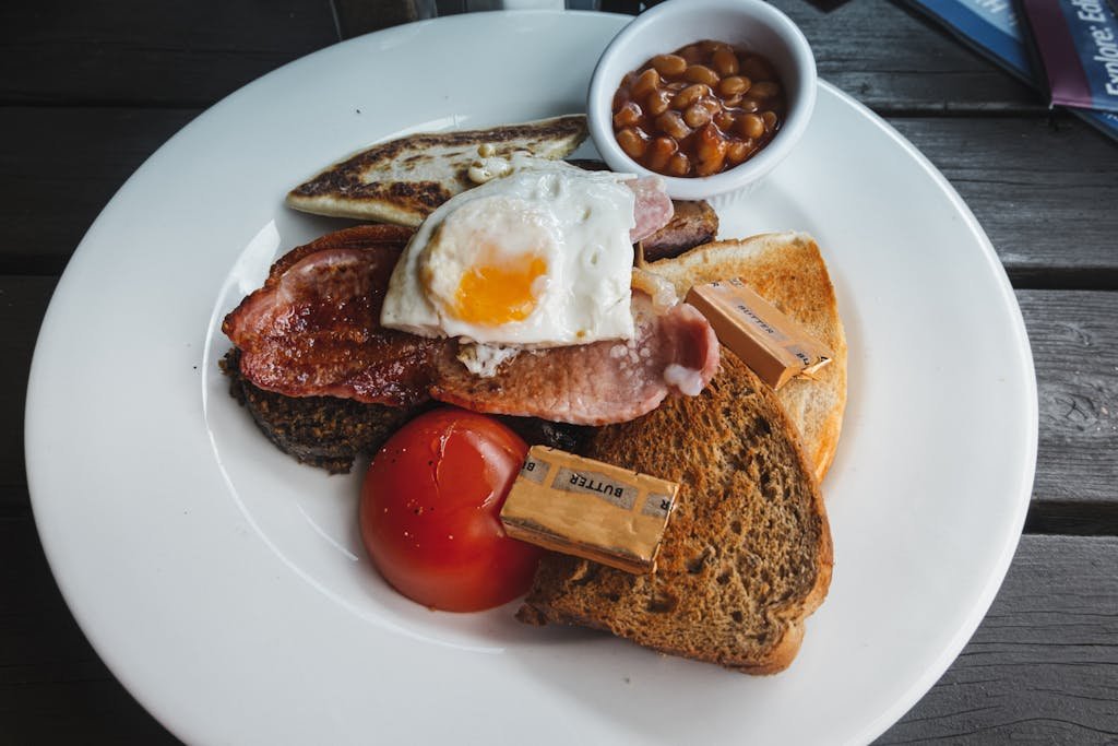 From above of toasted bread with fried bacon and egg served on white table with tomato and beans on wooden table