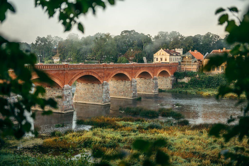 Historic red brick bridge over river in Kuldīga, Latvia surrounded by lush greenery.