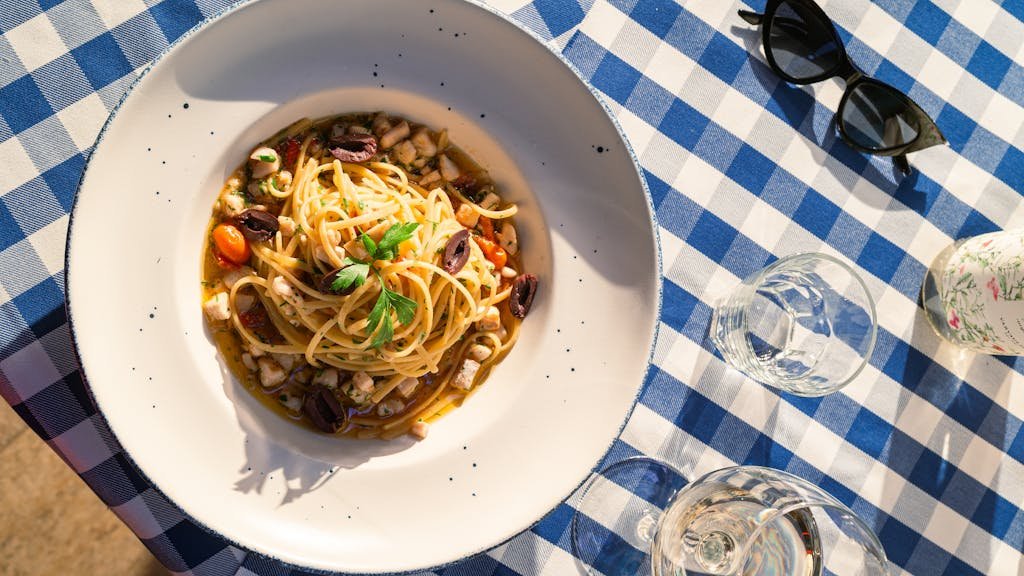 Is Malta Expensive? My Malta Budget Breakdown 2026 6 Top view of Italian pasta with parsley and olives on a blue checkered tablecloth with glasses in sunlight.