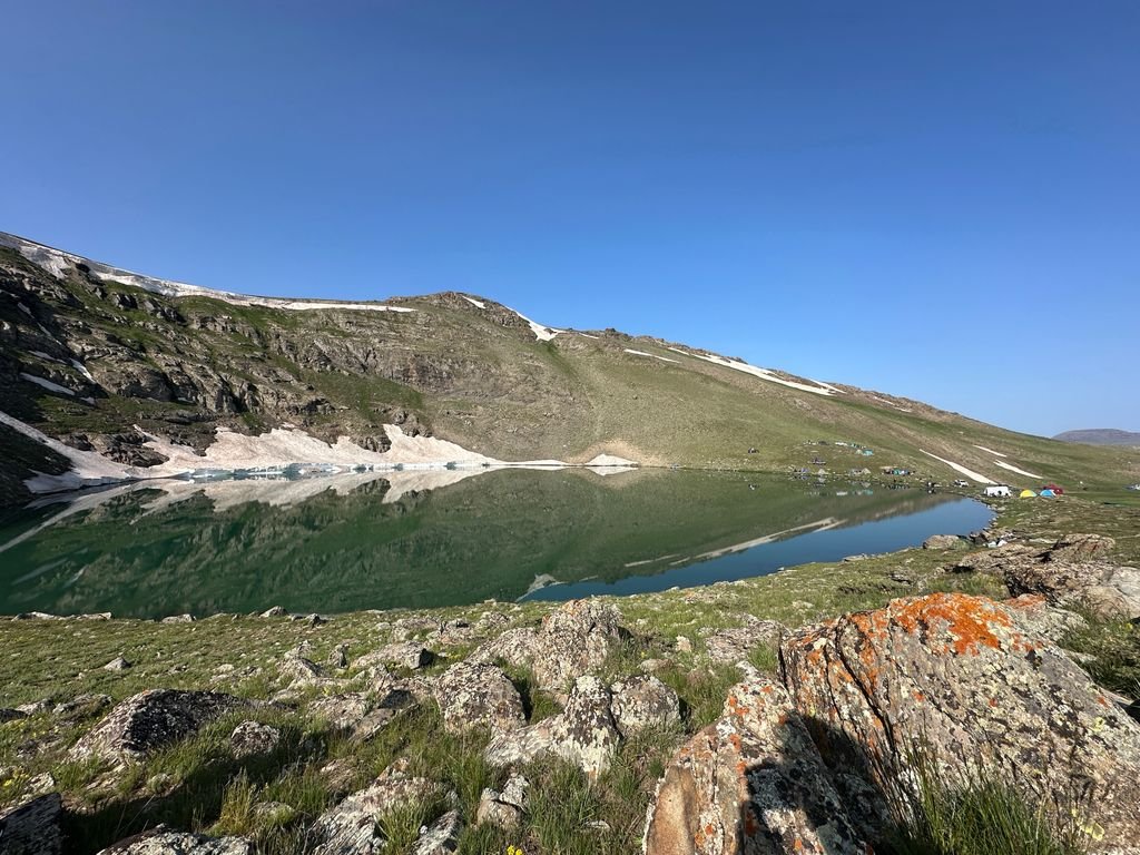 Dudipatsar Lake Trek: Pakistan's Most Beautiful Alpine Lake 7 Peaceful mountain lake reflecting rocky slopes and patches of snow under clear blue sky.