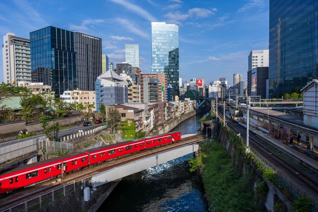 Red train crossing a bridge in Tokyo against a backdrop of modern skyscrapers and a clear blue sky.