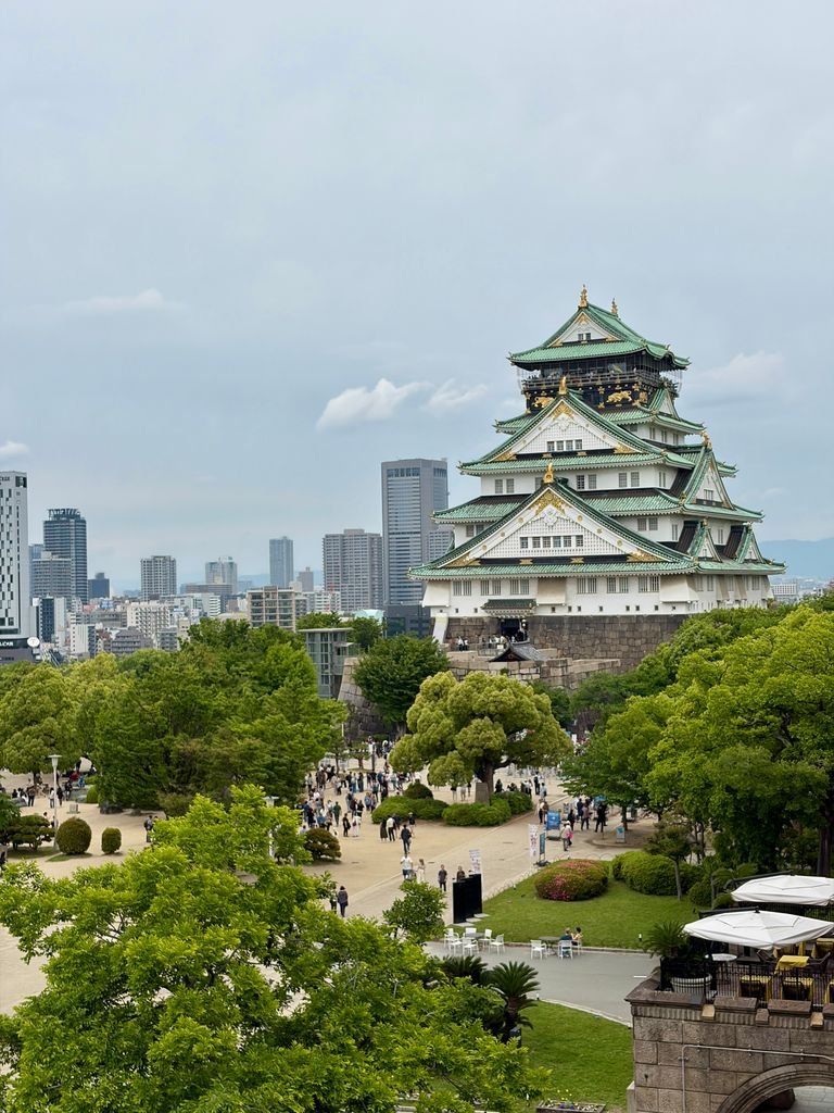 Scenic view of Osaka Castle surrounded by lush greenery and urban skyline on a cloudy spring day.