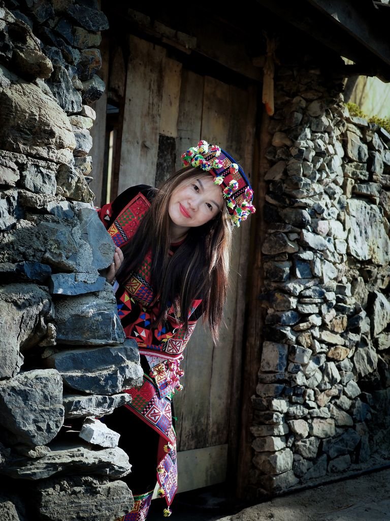 Kalash Valley Travel Guide 2026: Kalash Festivals, Culture & People 2 A young woman in vibrant traditional clothing poses by a rustic stone structure.