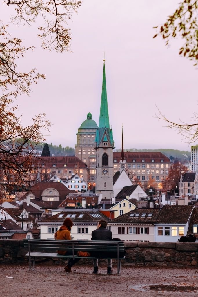 Couple sitting on a bench in Zurich with a skyline view including iconic church towers and urban landscape.